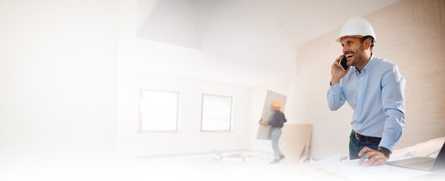 Smiling construction manager in hard hat talking on phone while reviewing blueprints at construction site with worker in background