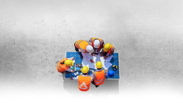 Aerial view of construction workers in hard hats and high-visibility safety vests gathered around blueprints on table at construction site