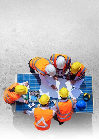 Aerial view of construction workers in hard hats and high-visibility safety vests gathered around blueprints on table at construction site