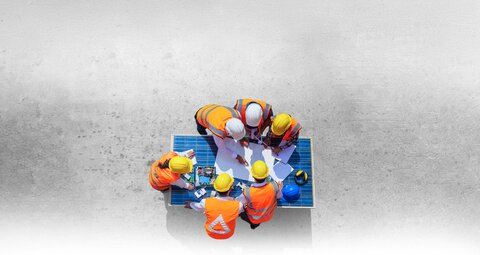 Aerial view of construction workers in hard hats and high-visibility safety vests gathered around blueprints on table at construction site
